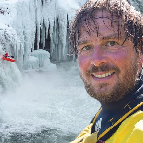 Im Vordergrund sieht man das Gesicht von Olaf Obsommer. Er hat nasse Haare, und lächelt in die Kamera. Im Hintergrund sieht man, wie eine Person im Kajak einen Wasserfall runterfährt. Der Wasserfall ist zum Großteil vereist.