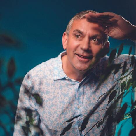 Man sieht Wladimir Kaminer, wie er durch eine Hecke oder einen Busch durchschaut. Sein Gesichtsausdruck ist neugierig, gespannt und vergnügt. Das Bild soll ihn beim Beobachten des "Geheimen Leben der Deutschen" zeigen.