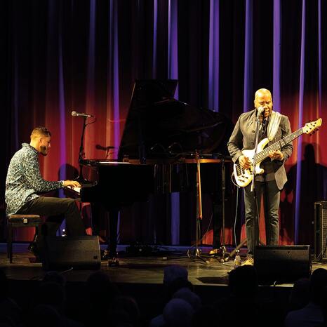 Auf der Bühne sind zwei Musiker zu sehen. Einer sitzt an einem Flügel und spielt. De andere spielt eine E-Gitarre und singt in ein Mikrofon. Die Lichtstimmung ist dunkel und bläulich gehalten.
