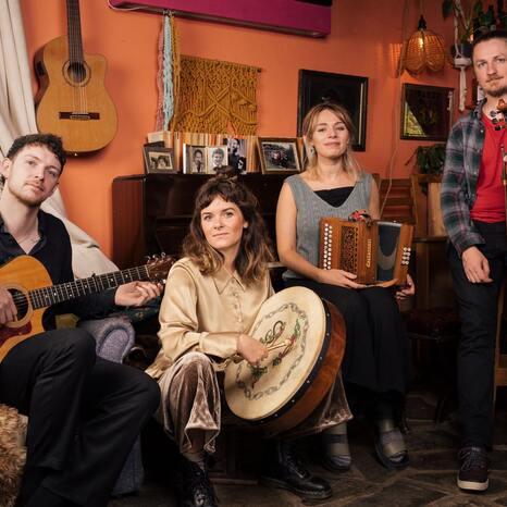 Die Band Eva Coyle Band besteht aus vier Personen. Sie sitzen auf dem Bild in einem gemütlichen Wohnzimmer und lächeln in die Kamera. Sie haben ihre Instrumente, eine akustische Gitarre, eine Trommel, ein Akkordeon und eine Fiedel bei sich.