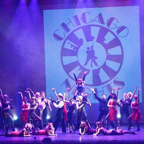 Das Odyssey Dance Theater steht auf der Bühne. Es sind 21 Menschen zu sehen. Alle tragen chice Outfits im zwanziger Jahre Look. Die Tanztruppe steht gerade in einem Standbild. Im Hintergrund hängt ein Banner mit der Aufschrift "Chicago Nights".