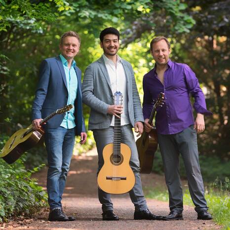 Das Bild zeigt die drei Musiker des Alegrias Guitar Trio. Sie stehen auf einem Weg, umgeben von grünen Bäumen. Sie halten ihre Gitarren und lachen in die Kamera.