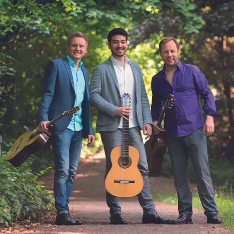 Das Bild zeigt die drei Musiker des Alegrias Guitar Trio. Sie stehen auf einem Weg, umgeben von grünen Bäumen. Sie halten ihre Gitarren und lachen in die Kamera.