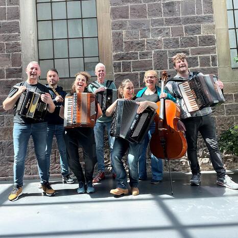 Eine Gruppe von sieben Personen ist zu sehen. Sie stehen vor einer Mauer aus Stein. Fünf von ihnen halten Akkordeons. Eine Person hält ein Chello. Alle lachen.